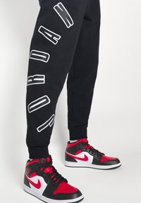 Pantalones de chándal negros con letras blancas a lo largo del lado y zapatillas altas rojas y negras con detalles en blanco y cordones negros.