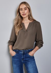 Olivgrön blus med V-ringning och rynkade ärmar, i kombination med blå jeans. Mjuk tyg, lös passform, minimalistisk design.
