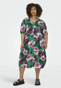 Blumiges Midi-Kleid mit grünem Grundton, das große pinke und weiße Blumen zeigt, kurze Puffärmel, einen V-Ausschnitt und Knopfdetails auf der Vorderseite hat.