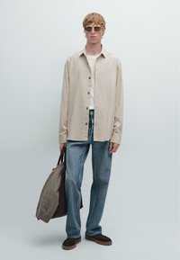 Ljust beige skjorta med svarta knappar, vit t-shirt under, blå jeans, bruna loafers och en grå tygväska.