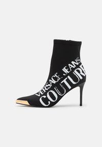 Czarne botki za kostkę na wysokim obcasie, z białym napisem "VERSACE JEANS COUTURE" oraz metalicznie złotym czubkiem na spiczastym nosku. Wykonane z elastycznego materiału.