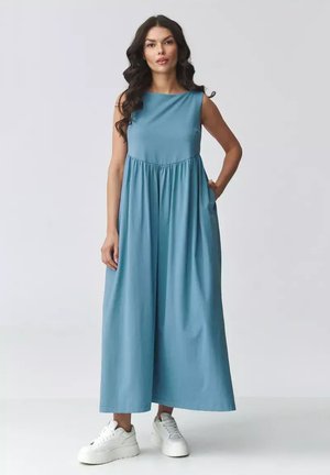 Kobieta o długich ciemnych włosach, ubrana w niebieską, luźną, długą sukienkę bez rękawów i białe trampki, stojąca z jedną ręką w kieszeni.
