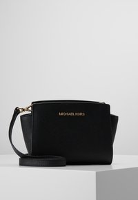 Černá kožená kabelka se strukturovaným designem, zlatými kovovými detaily a odnímatelným popruhem. Na přední straně se nachází logo Michael Kors.