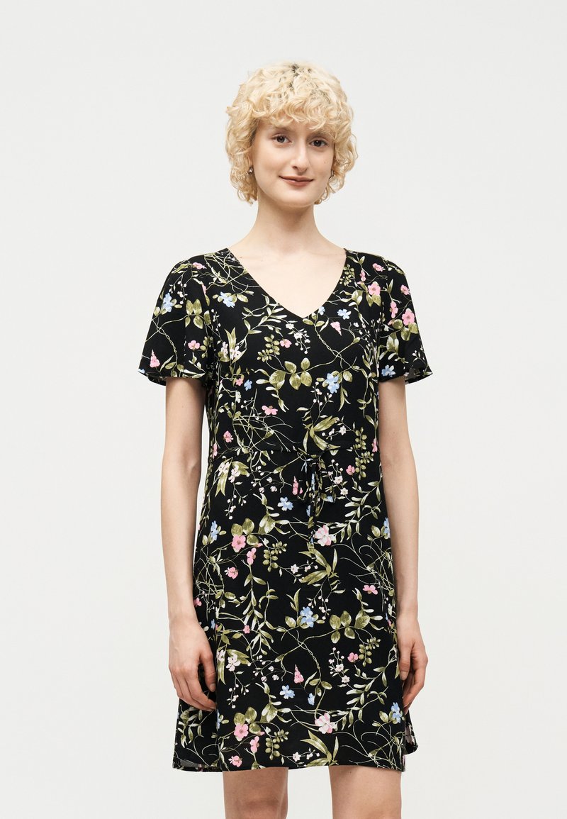 Νεαρό άτομο με κοντά, σγουρά ξανθά μαλλιά, που φοράει μαύρο φόρεμα με φλοράλ σχέδιο και κοντά μανίκια, στέκεται μπροστά σε ένα απλό φόντο.