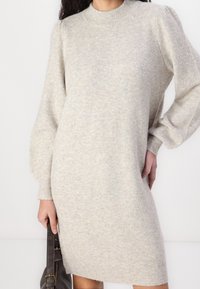 Femme portant une robe pull tricotée beige clair avec des manches bouffantes, tenant un sac à main en cuir marron foncé à côté d'elle.