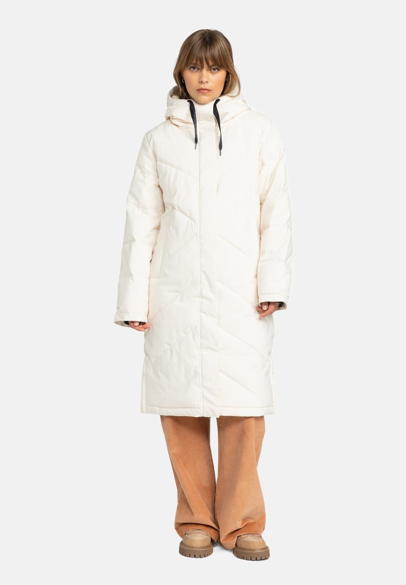 Long manteau matelassé en blanc cassé avec capuche, présentant un design matelassé, des cordons de serrage noirs et une silhouette spacieuse, associé à un pantalon large beige.