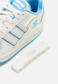 Vita lädersneakers med blå accenter, som har tre texturerade ränder, en vadderad krage och ett avtagbart bälte. Adidas-logotypen visas.