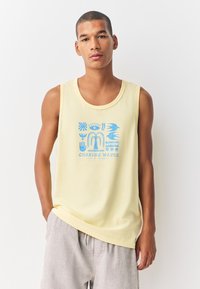 Gaiši dzeltena bezrokas krekla virskrekls ar zilu grafisko dizainu, kurā ir stilizētas formas un teksts "CHASING WAVES", kas centrēts priekšpusē.