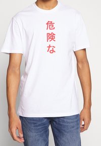 Bílé tričko s červenými vertikálními japonskými znaky, které nosí člověk v modrých džínách, stojící na jednobarevném pozadí.