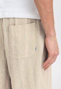Un pantalon en tissu côtelé beige clair présente une poche latérale, avec une petite étiquette pour le branding. Le matériau semble doux avec une finition texturée.