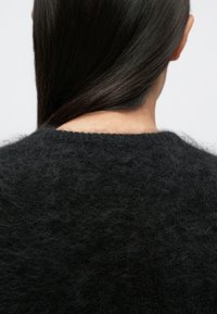 Pulover țesut negru cu o textură moale și pufoasă. Are un decolteu rotund și o croială ușor lejeră, arătând partea din spate a piesei vestimentare și părul.