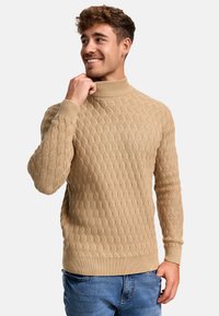 Pull en tricot beige à col montant, avec un motif de vagues texturé et des poignets et un ourlet côtelés. Assorti avec un jean bleu.