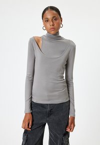 Helles graues Langarmshirt mit Stehkragen, das ein einzigartiges Ausschnittdesign an einer Schulter aufweist und aus einem glatten, dehnbaren Material gefertigt ist.