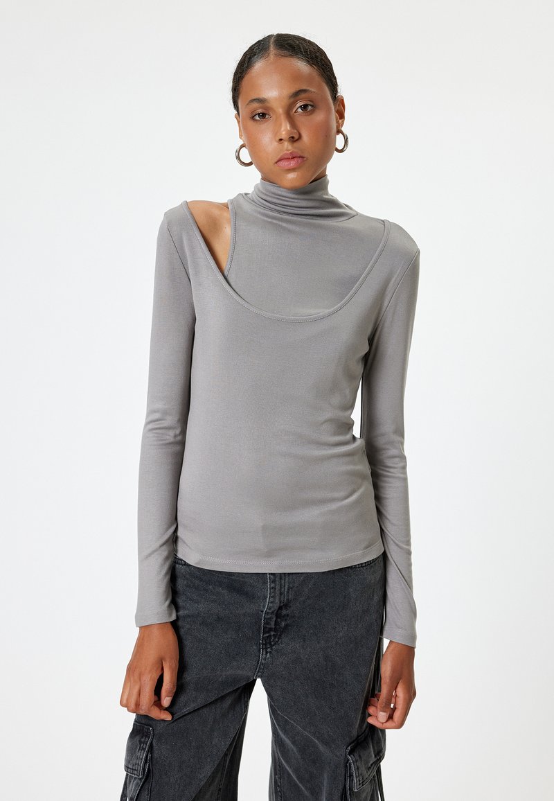 Helles graues Langarmshirt mit Stehkragen, das ein einzigartiges Ausschnittdesign an einer Schulter aufweist und aus einem glatten, dehnbaren Material gefertigt ist.