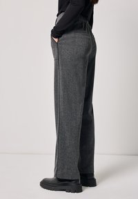 Wijde broek van donkergrijs stof met een subtiele textuur, voorzien van een elastische tailleband en zijzakken, gecombineerd met zwarte schoeisel.
