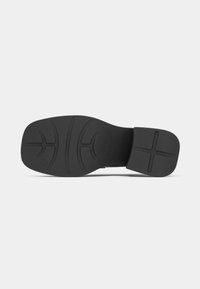 Semelle d'une chaussure noire à bout carré avec des motifs texturés sur la semelle et un petit talon carré, sur un fond clair uni.