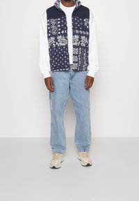 Man som bär en väst med svart och vit paisleymönster över en vit långärmad skjorta, ljusblå jeans och beige sneakers, stående framför en enfärgad bakgrund.