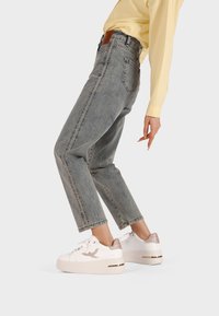 Persona che indossa jeans blu sbiaditi e sneakers con piattaforma bianche con dettagli in glitter rosa oro, in piedi contro uno sfondo chiaro e uniforme.