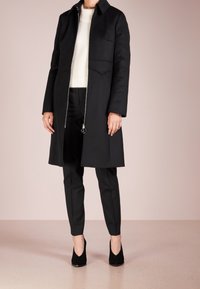 Cappotto di lana nera con colletto alto, chiusura con zip e due tasche frontali, abbinato a pantaloni neri e scarpe con tacco alto.