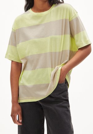 Henkilö pukeutuneena löysään t-paitaan, jossa leveät vaakasuorat vaaleankeltaiset ja beiget raidat, toinen käsi mustien housujen taskussa.