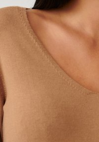 Haut en tricot couleur fauve avec un décolleté en V profond, une texture lisse et des coutures visibles le long de l'encolure. Tissu doux avec une légère brillance.