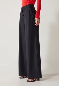 Pantalons large en tissu noir, avec une taille haute, des boutons sur le côté et une texture lisse, associés à des sandales à talons hauts noires.