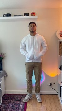 Sudadera blanca con cremallera, pantalones cargo beige, zapatillas grises. Incluye bolsillos tipo canguro. De pie sobre un suelo de madera, con luces coloridas de fondo.