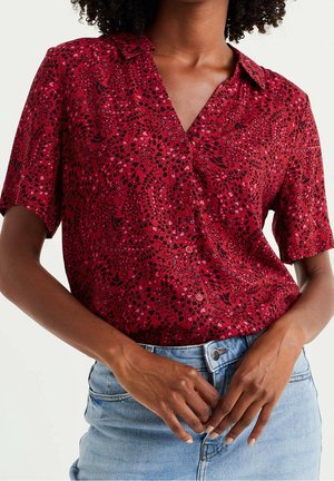 Czerwona bluzka z krótkim rękawem, zdobiona różowym i czarnym wzorem kwiatowym, z kołnierzykiem i guzikami z przodu. Połączona z dżinsowymi szortami.