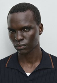Polo shirt nero a maniche corte con colletto marcato e dettagli in cucitura arancio, indossato sopra una maglietta bianca. Aspetto liscio e texturizzato.