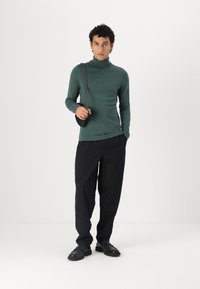 Groene coltrui, slim-fit zwarte broek en zwarte veterschoenen. Model poseert met een kleine crossbody tas. Glad textuur.
