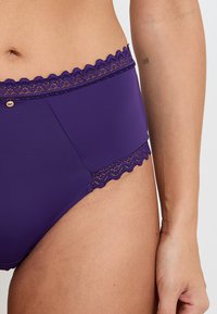 Parte inferiore di bikini viola con una texture liscia, orlo in pizzo in vita e un piccolo accento dorato. Presenta un design a vita alta.
