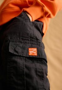 Čierne cargo nohavice majú textúrovanú tkaninu s viditeľnou oranžovou nášivkou „Super Dry“ na boku; skombinované s oranžovou košeľou.