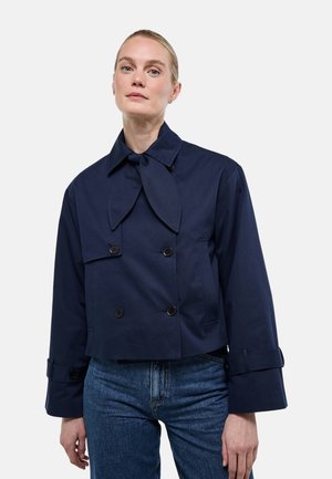 Femme portant une veste courte bleu marine à double boutonnage, avec un grand nœud au col et des brides aux poignets, associée à un jean bleu.
