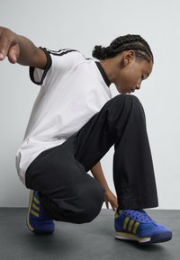 Blue and yellow Adidas shoes, black loose pants, and a white t-shirt with black accents, crouching on a gray surface, focused on the shoes.