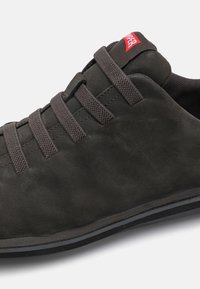 Tumehall suede jalanõud tasa talla ja elastsete paeltega, millel on väike punane logo keele peal. Sile tekstuur ja ümar kuju.