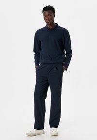 Tummansininen pitkähihaiset polo-paita, jossa on kaulus, yhdistettynä tummansinisiin housuihin. Malli seisoo valkoisissa lenkkareissa, esitellen yksinkertaista ja yhtenäistä ilmettä.