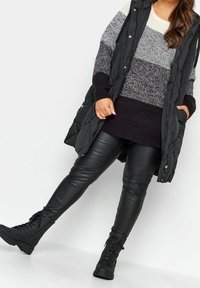 Svart vadderad väst över en färgblockerad tröja med grå och krämfärgade ränder, tillsammans med svarta byxor i konstläder och snörade ankelskor.