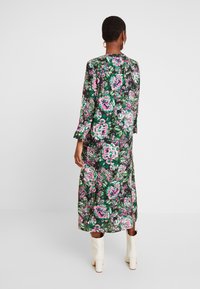 Mujer con el cabello corto que lleva un vestido negro de manga larga con estampado floral en tonos rosa y verde, botines blancos, de pie contra un fondo blanco.
