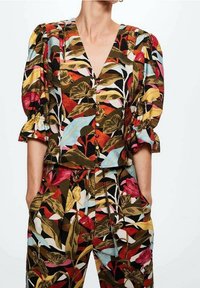 Donna che indossa un top floreale abbinato con maniche a sbuffo e pantaloni caratterizzati da grandi foglie e fiori in tonalità di rosso, giallo, blu e marrone.