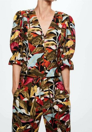 Donna che indossa un top floreale abbinato con maniche a sbuffo e pantaloni caratterizzati da grandi foglie e fiori in tonalità di rosso, giallo, blu e marrone.