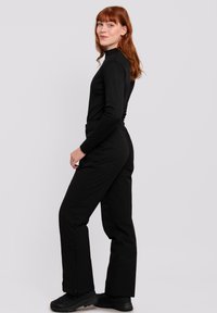 Schwarzes Langarmshirt und passende schwarze Hose. Die Hose hat Reißverschlüsse an den Knöcheln und das Outfit hat eine figuroptimierende Silhouette.