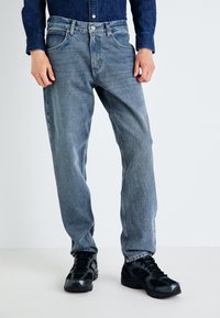 Jeans de mezclilla azul claro con un corte recto, que cuentan con cinco bolsillos, un cierre frontal con botón y un ligero desgastado. Llevados con zapatillas negras.