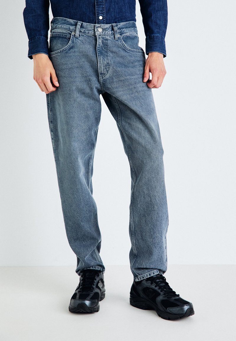 Jeans de mezclilla azul claro con un corte recto, que cuentan con cinco bolsillos, un cierre frontal con botón y un ligero desgastado. Llevados con zapatillas negras.