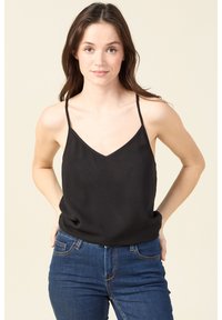 Schwarzes Spaghetti-Trägertop mit V-Ausschnitt und glatter Textur, kombiniert mit dunkelblauen, eng anliegenden Jeans in einer lässigen Pose.