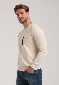 Sudadera beige con cuello redondo, mangas largas y un bolsillo en el pecho izquierdo con una cremallera negra. Textura suave, diseño casual y estilo ajustado.