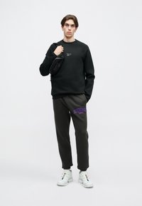 Joven de pie con sudadera negra de Reebok, pantalones de chándal oscuros con el logo morado, zapatillas blancas y sosteniendo un bolso cruzado negro.