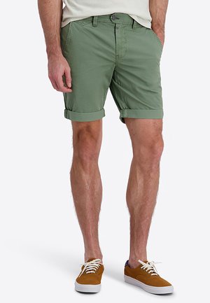 Mann trägt hochgekrempelte olivgrüne Shorts, weißes T-Shirt und braune Schnürsneaker und steht mit den Händen in den Taschen.