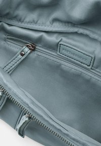 Interno di una borsa di tessuto verde che mostra una texture liscia, una tasca con zip e un'etichetta. Le caratteristiche includono hardware metallizzato e dettagli di cucitura.