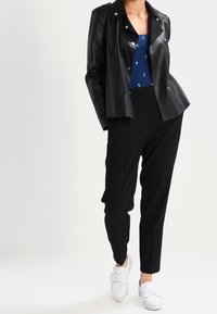 Mujer con chaqueta de piel sintética negra, blusa azul con estampado, pantalones negros ajustados y zapatillas blancas sin cordones con lazos.