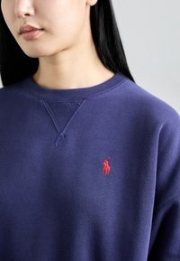 Tmavofialová mikina s okrúhlym výstrihom a rebrovanými manžetami, s malým červeným vyšitým logom na ľavej časti hrude. Mäkká textúra.